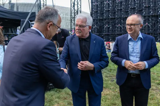 Trzech mężczyzn w garniturach w trakcie powitania na wydarzeniu plenerowym.