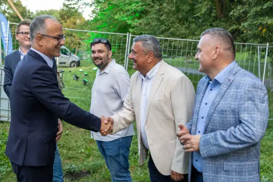 Grupa mężczyzn w formalnych strojach, uściski dłoni i rozmowy na tle zieleni.