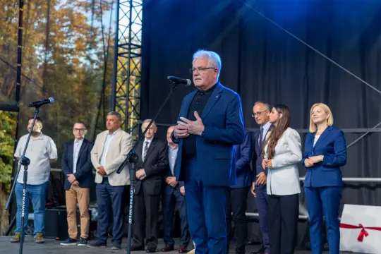 Mężczyzna przemawia do zgromadzonej publiczności, w tle grupa osób na scenie.