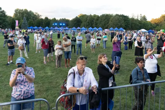 Tłum ludzi na festiwalu, niektórzy trzymają telefony, w tle namioty i scena.