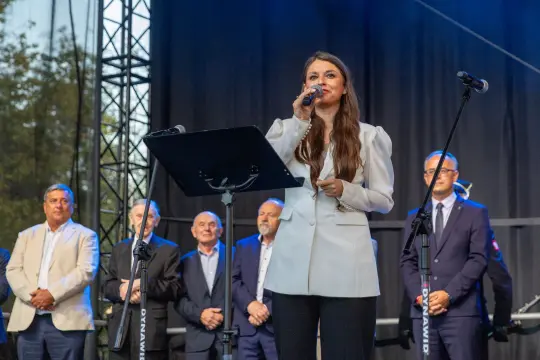 Kobieta przemawia do publiczności, w tle stoją mężczyźni w garniturach.