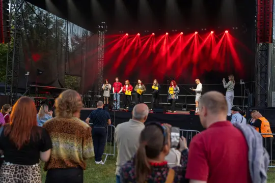 Grupa osób na scenie podczas wydarzenia, w tle czerwone światła. Publiczność z zainteresowaniem obserwuje.