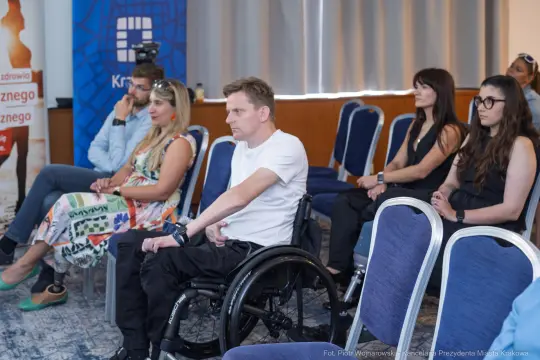 Grupa osób siedzących na krzesłach, w tym jedna na wózku inwalidzkim.