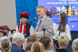Mężczyzna przemawia na inauguracji roku szkolnego, obok niego uczniowie w tradycyjnych strojach.