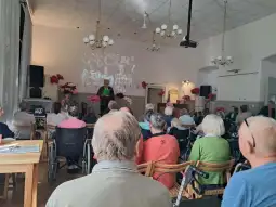 Spotkanie seniorów w sali z prezentacją na ekranie i dekoracjami.