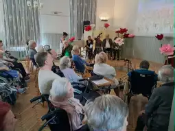 Spotkanie seniorów z występem artystycznym w sali z dekoracjami kwiatowymi.