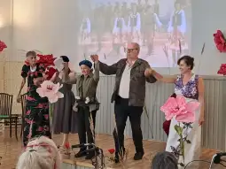 Grupa osób na scenie w trakcie występu, w tle projekcja i duże kwiaty.