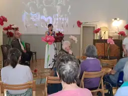 Kobieta przemawia na tle ekranu, w sali z kwiatami i słuchającą publicznością.