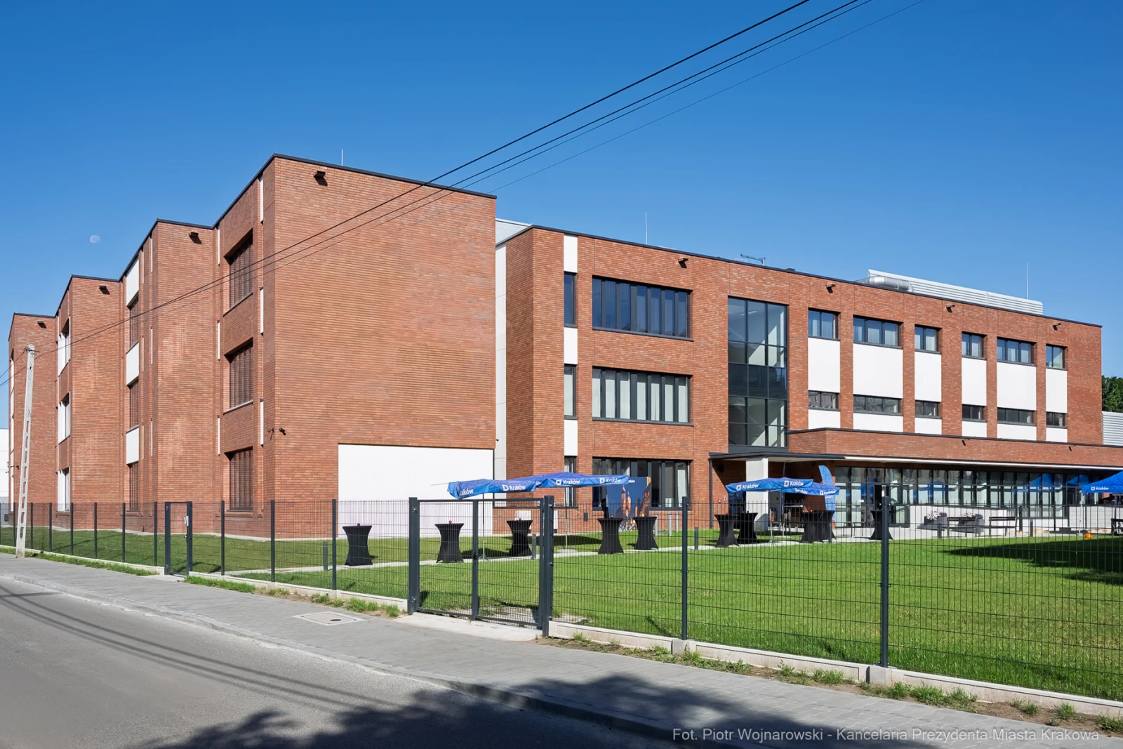 Nowoczesny budynek z cegły, otoczony zielenią i ogrodzeniem, z tarasem i parasolami.  Autor: Piotr Wojnarowski