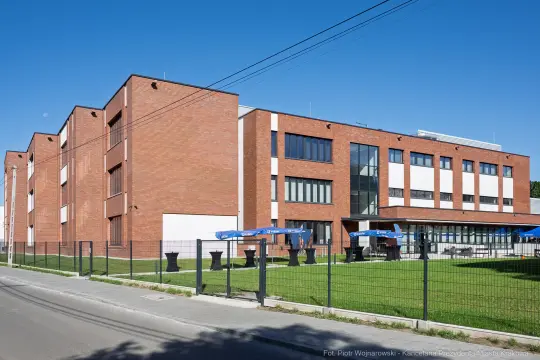 Nowoczesny budynek z cegły, otoczony zielenią i ogrodzeniem, z tarasem i parasolami.