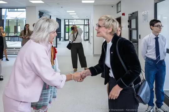 Spotkanie dwóch kobiet, jedna z nich uścisnęła dłoń drugiej. W tle inni uczestnicy wydarzenia.