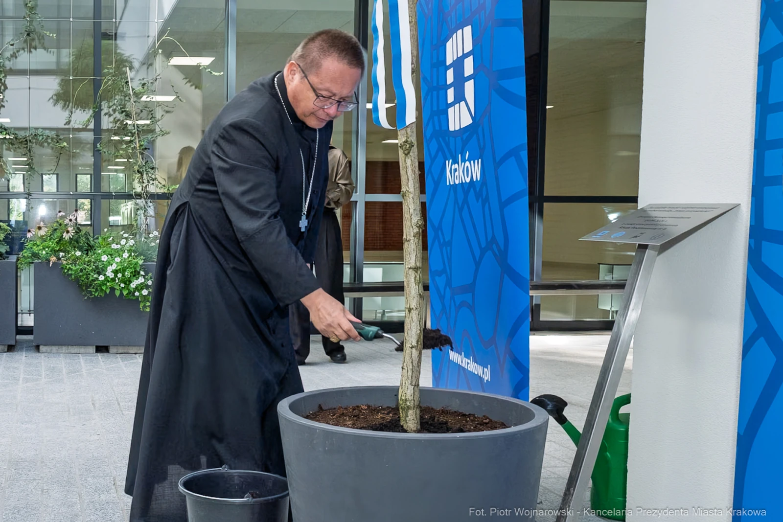 Ksiądz sadzi drzewo w donicy w otoczeniu nowoczesnego budynku.  Autor: Piotr Wojnarowski