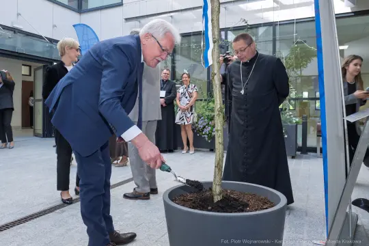 Sadzenie drzewa przez mężczyznę w garniturze, w tle inni uczestnicy wydarzenia.