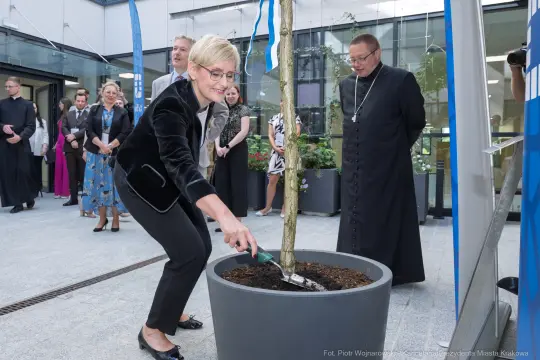 Sadzenie drzewa przez kobietę w eleganckim stroju, w towarzystwie innych osób.