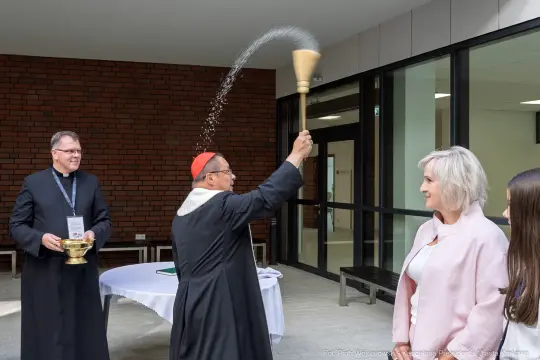 Kardynał błogosławi wodą, obok stoją osoby w strojach liturgicznych i cywilnych.