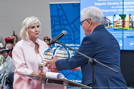 Kobieta wręcza mężczyźnie stos książek podczas inauguracji roku szkolnego.