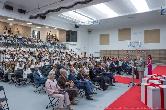 Uroczystość w sali gimnastycznej z uczestnikami siedzącymi na widowni i mówcą na podium.