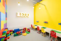 Pokój zabaw z kolorowymi klockami LEGO, żółtymi ścianami i stolikami dla dzieci.