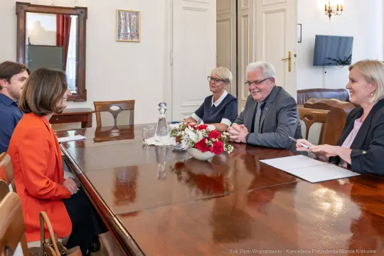 Spotkanie przy stole, cztery osoby rozmawiają, na stole kwiaty i woda.