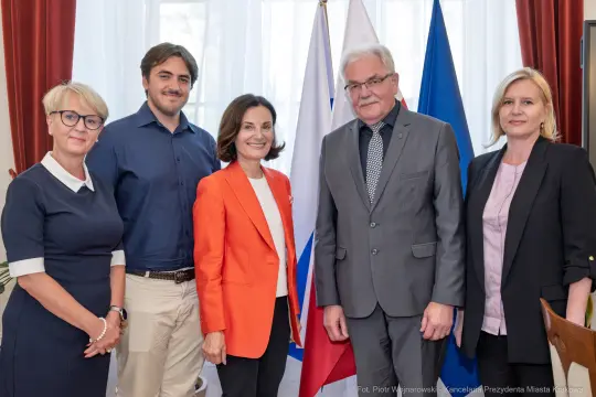 Cztery osoby w formalnych strojach stoją obok siebie, w tle flagi Polski i Unii Europejskiej.