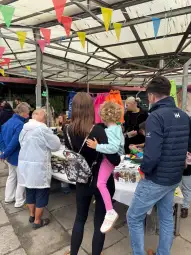 Targ z ludźmi, straganami i kolorowymi flagami, dziecko na rękach matki.