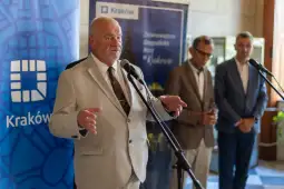 Mężczyzna w jasnym garniturze przemawia do mikrofonu, w tle inni mężczyźni i banery promocyjne.