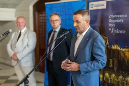 Mężczyzna w garniturze przemawia do mikrofonu, w tle dwóch mężczyzn i baner promujący Kraków.