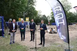 Konferencja prasowa z udziałem mężczyzn w garniturach i flagą. W tle sprzęt budowlany.