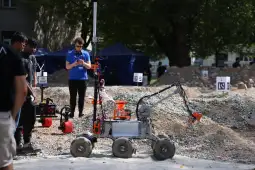 Robot z ramieniem i kołami, uczestniczący w zawodach w terenie. W tle ludzie i namioty.