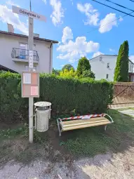 Przystanek autobusowy z ławką i koszem na śmieci, otoczony zielenią.