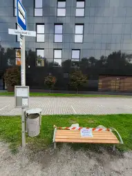Ławka przystankowa z tabliczką informacyjną, obok kosz na śmieci i znak przystanku.