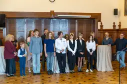 Grupa osób w formalnych strojach, stojących w sali z drewnianymi ścianami.