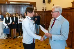 Wręczenie dyplomu przez mężczyznę w garniturze młodemu mężczyźnie w białej koszuli. W tle grupa osób.