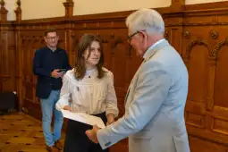 Wręczenie dyplomu młodej kobiecie przez starszego mężczyznę w eleganckim wnętrzu.
