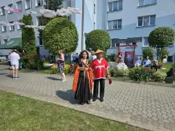 Kobieta w czarnej sukni i mężczyzna w tradycyjnym stroju na tle ogrodu i budynków.