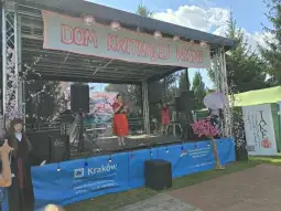 Kobieta w czerwonej sukience śpiewa na scenie, obok dekoracje i instrumenty muzyczne.