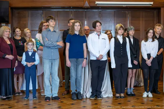 Grupa osób w formalnych strojach, stojących w sali z drewnianymi panelami.