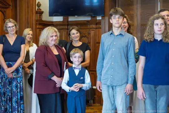 Grupa osób w eleganckich strojach, w tym chłopiec w muszce, w sali z drewnianymi elementami.