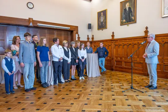 Grupa ludzi w eleganckich strojach podczas uroczystości w sali z drewnianymi panelami.