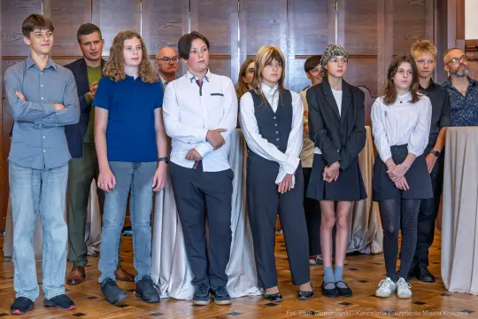 Grupa młodych ludzi w formalnych strojach, stojących w rządku na tle drewnianej ściany.