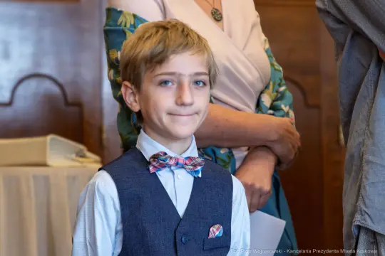 Chłopiec w eleganckim stroju z muszką, uśmiechający się w towarzystwie dorosłych.