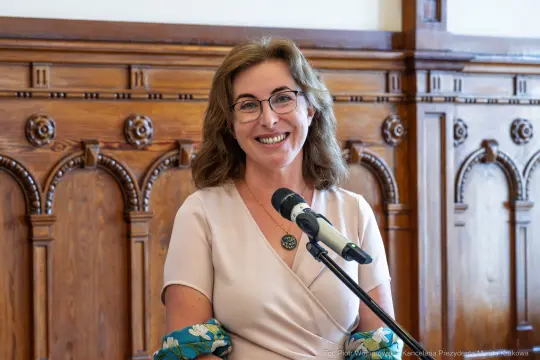 Kobieta z okularami uśmiecha się, trzymając mikrofon w eleganckim wnętrzu.
