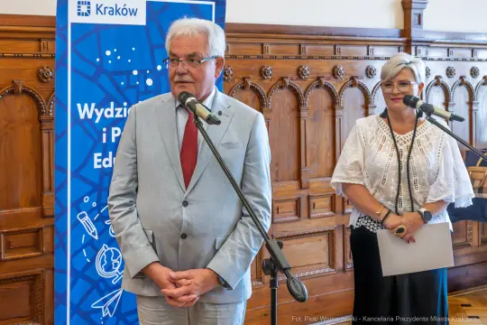 Mężczyzna w jasnym garniturze i kobieta w białej bluzce przy mikrofonach podczas wydarzenia w Krakowie.