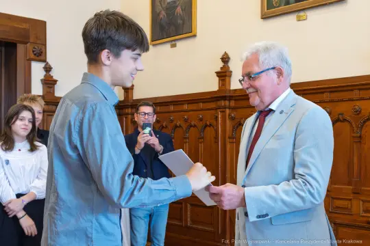 Młody mężczyzna wręcza dokument starszemu mężczyźnie w eleganckim otoczeniu.