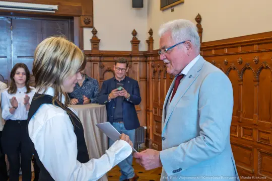Wręczenie dyplomu przez mężczyznę w garniturze młodej osobie w białej koszuli. W tle grupa osób.