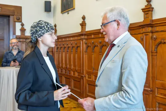 Młoda kobieta w okularach i czapce rozmawia z mężczyzną w garniturze w eleganckim wnętrzu.