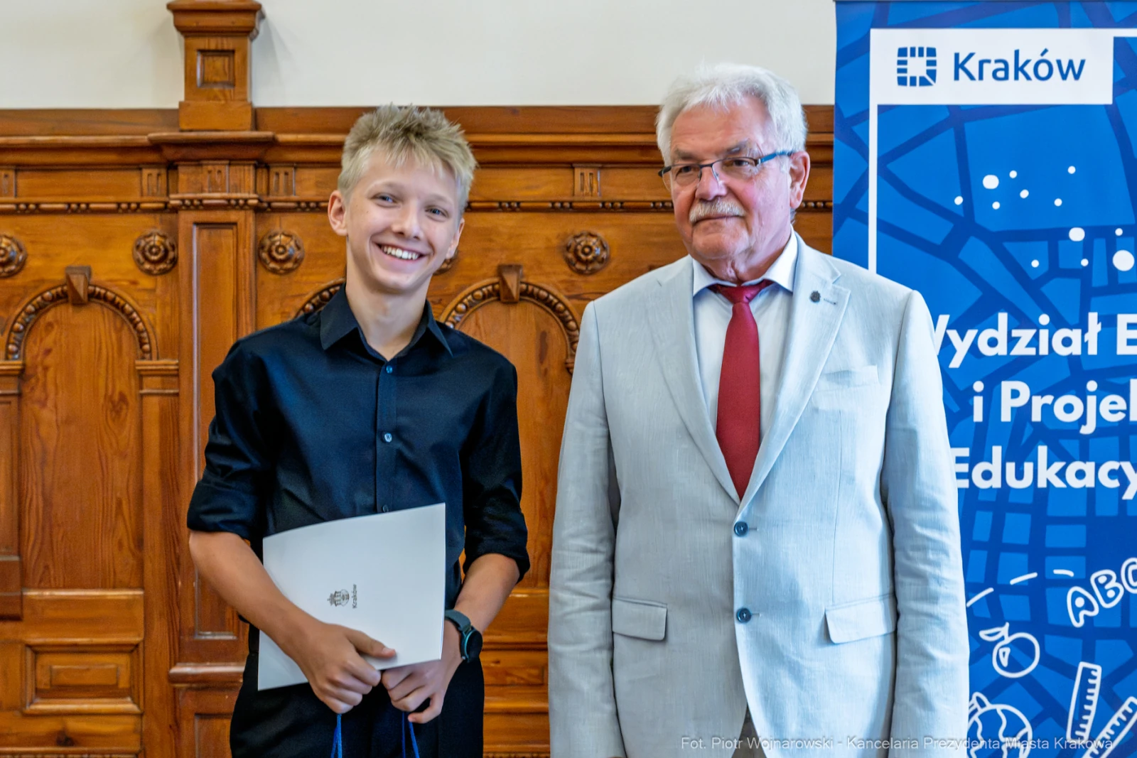 Chłopiec z dyplomem i mężczyzna w garniturze, obaj uśmiechnięci, w sali z drewnianymi elementami.  Autor: Piotr Wojnarowski