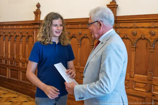 Młody chłopak odbiera list od starszego mężczyzny w eleganckim otoczeniu.