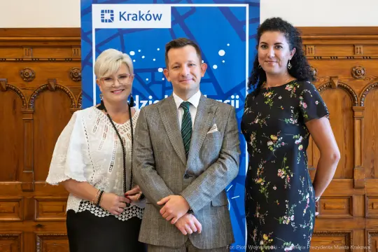 Trzy osoby pozujące do zdjęcia przed niebieskim tłem z logo Krakowa.