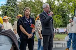 Mężczyzna z mikrofonem przemawia obok kobiety, w tle grupa ludzi w jasnych ubraniach.